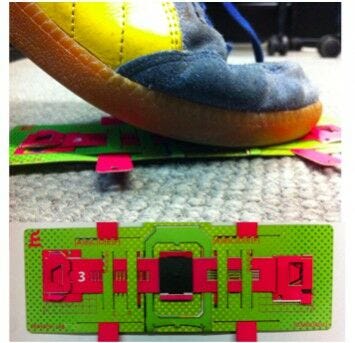 foot on FoldScope green and red paper foldscope on ground with shoe