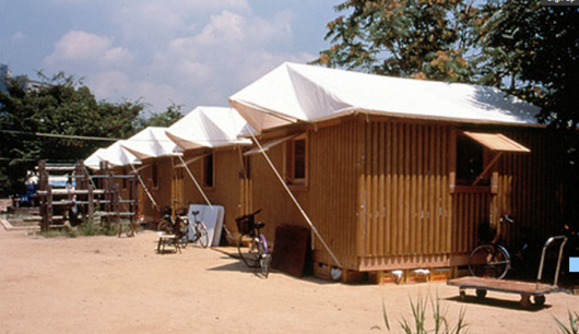 row of brown paper log houses with white tent roofs on dirt surrounded by trees