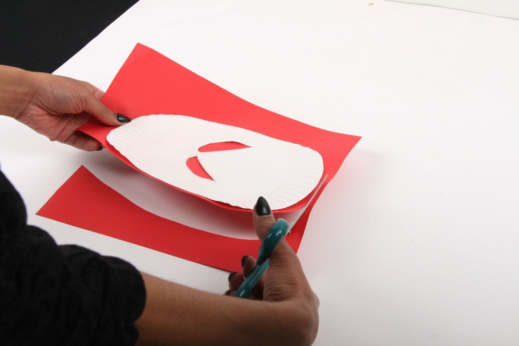 hands cutting around white paper plate on red paper with scissors