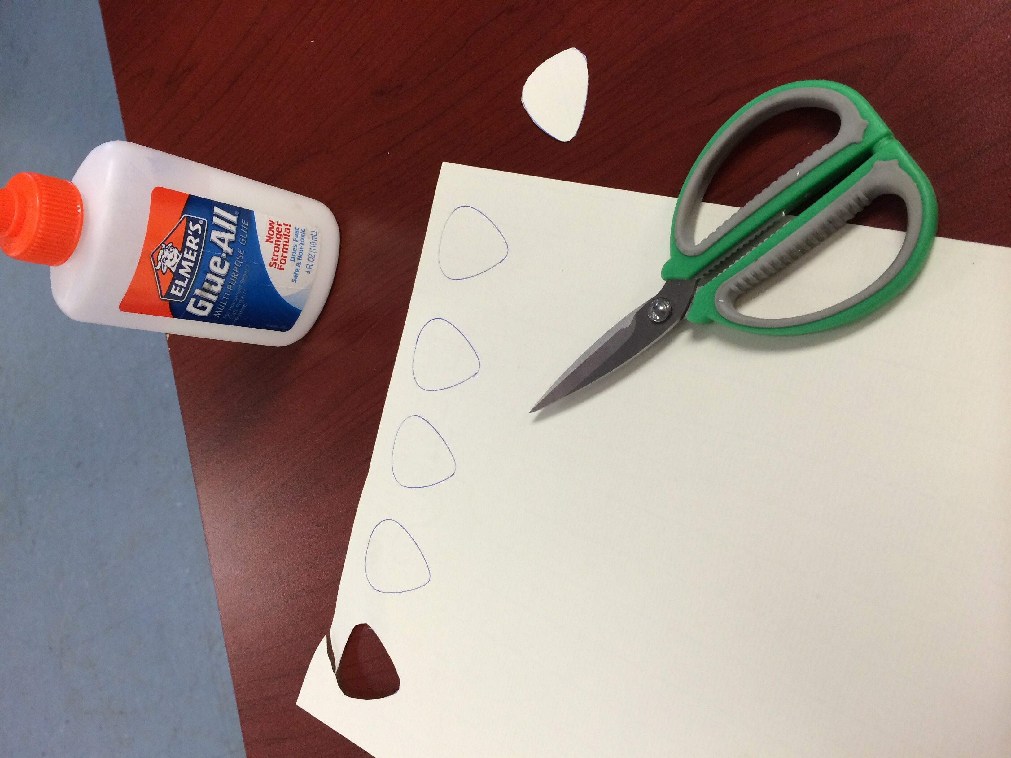 sheet of white cardstock with traced outlines of guitar picks on a desk with scissors and glue