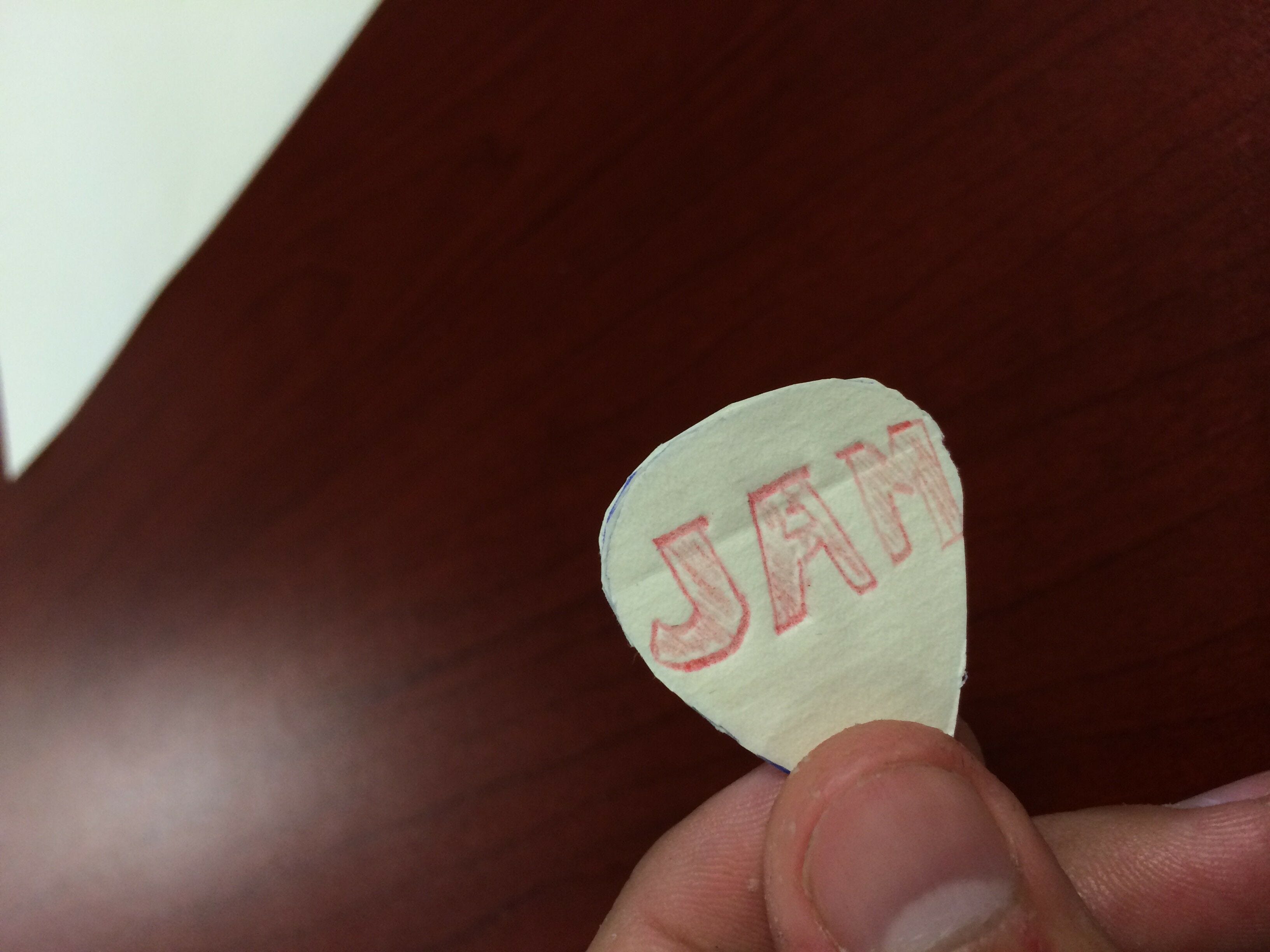 hand holding white paper guitar pick with red jam logo