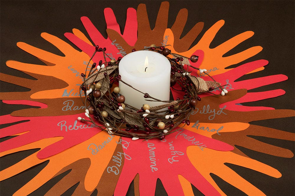 Thanksgiving centerpiece consisting of hand cutouts of fall-colored construction paper arranged in a circle with names inscribed on them in silver, and a white candle surrounded by a small wreath in the center